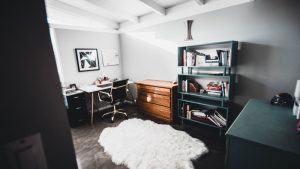 Small room with desk and chair in the corner next to a dresser and bookshelf with a white rug on the floor